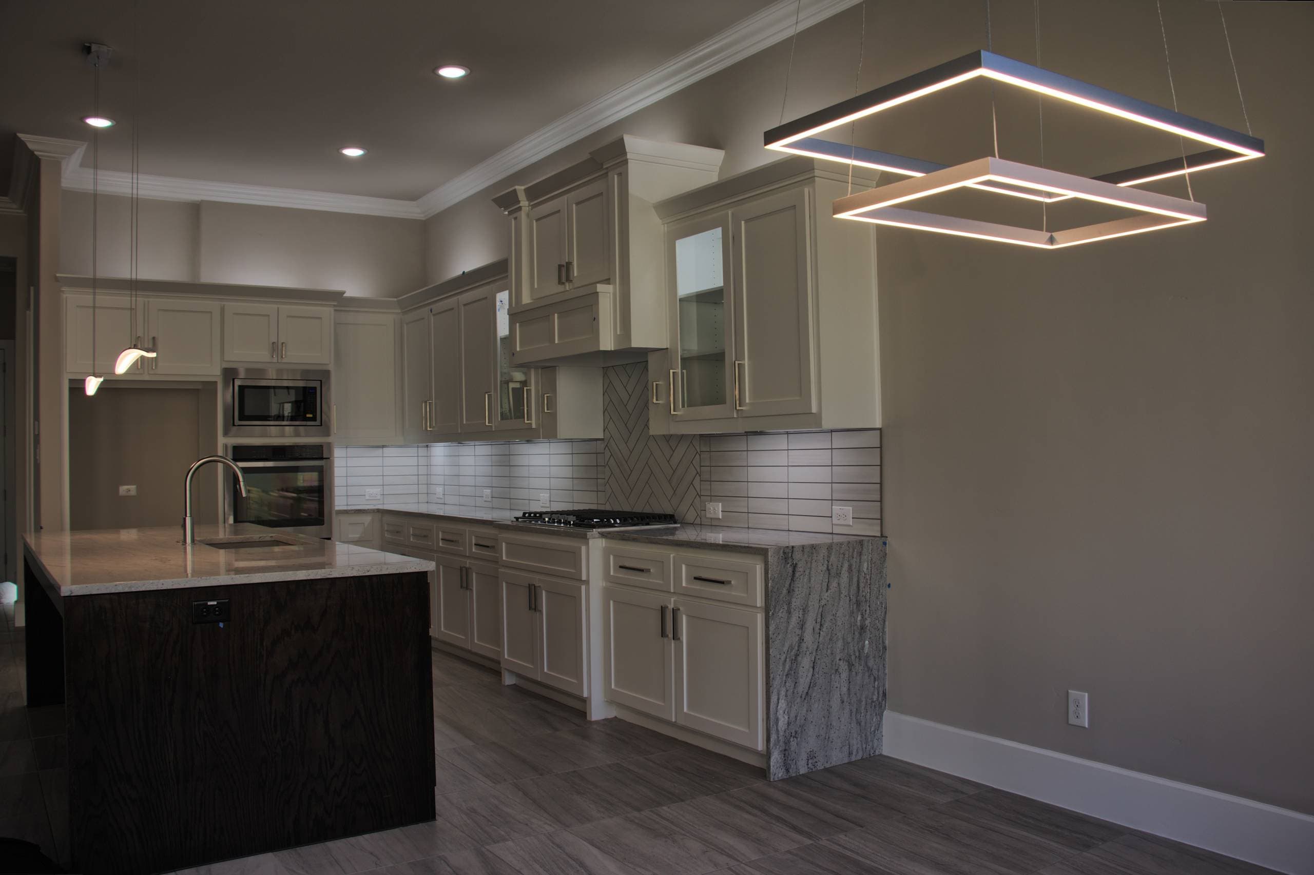 Two-tier square LED chandelier beside a kitchen with light gray shaker cabinets, granite waterfall counter, dark oak island, and herringbone tile accent
