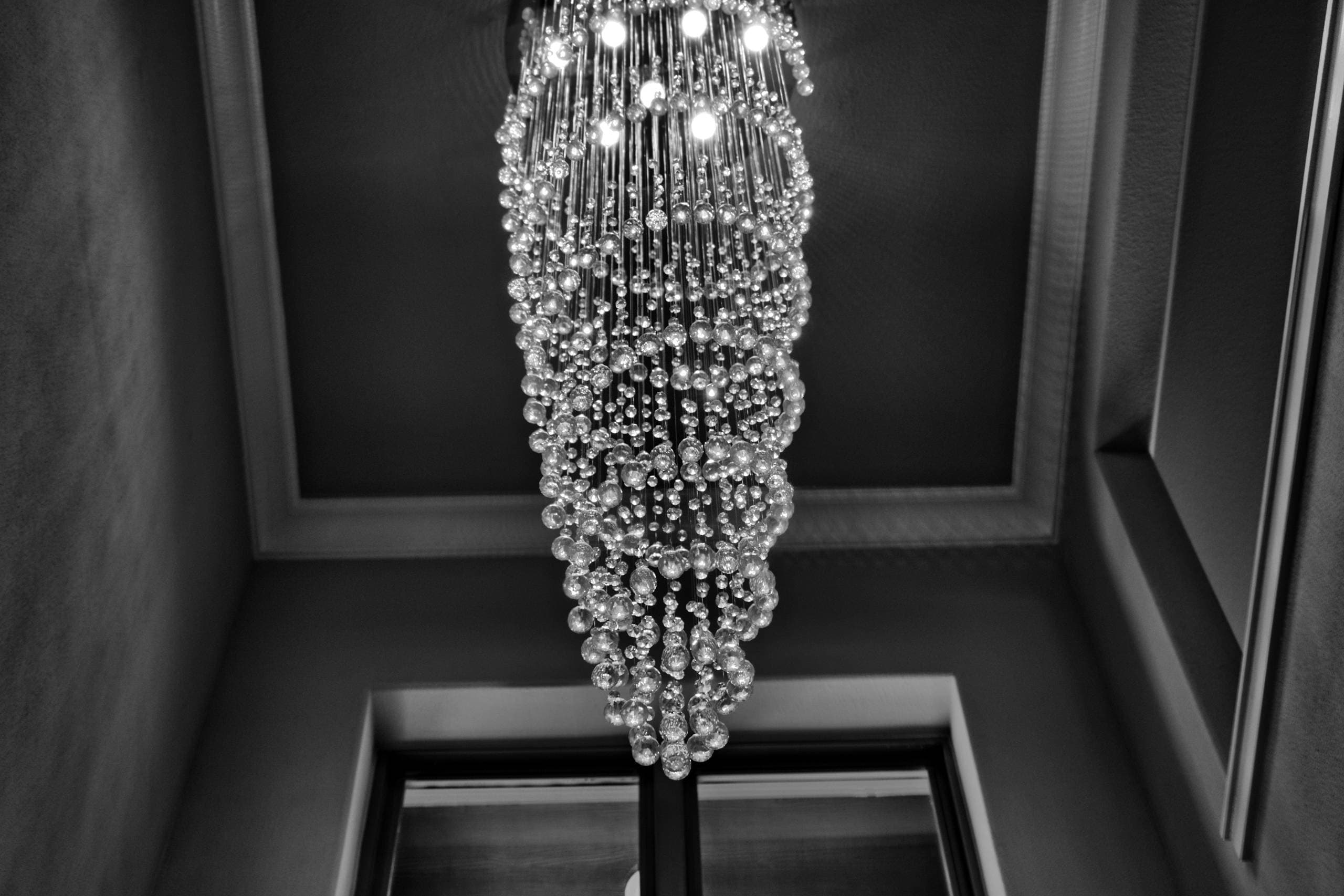 Black-and-white upward view of a tall spiral crystal raindrop chandelier hanging from a trimmed ceiling recess above a stairwell window
