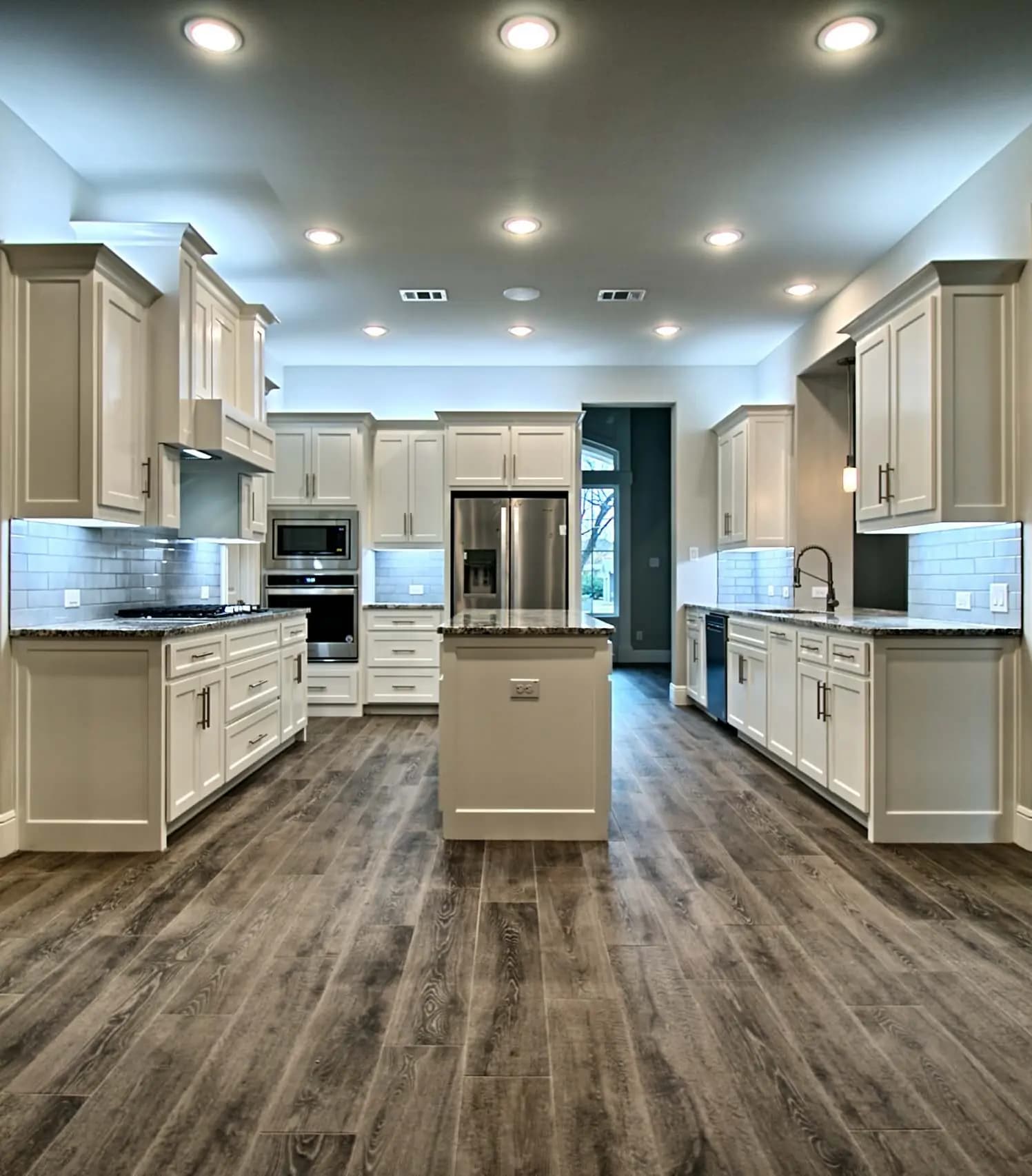 Bright, spacious kitchen with white cabinetry, stainless steel appliances, granite countertops, subway tile backsplash, and wide plank wood flooring.