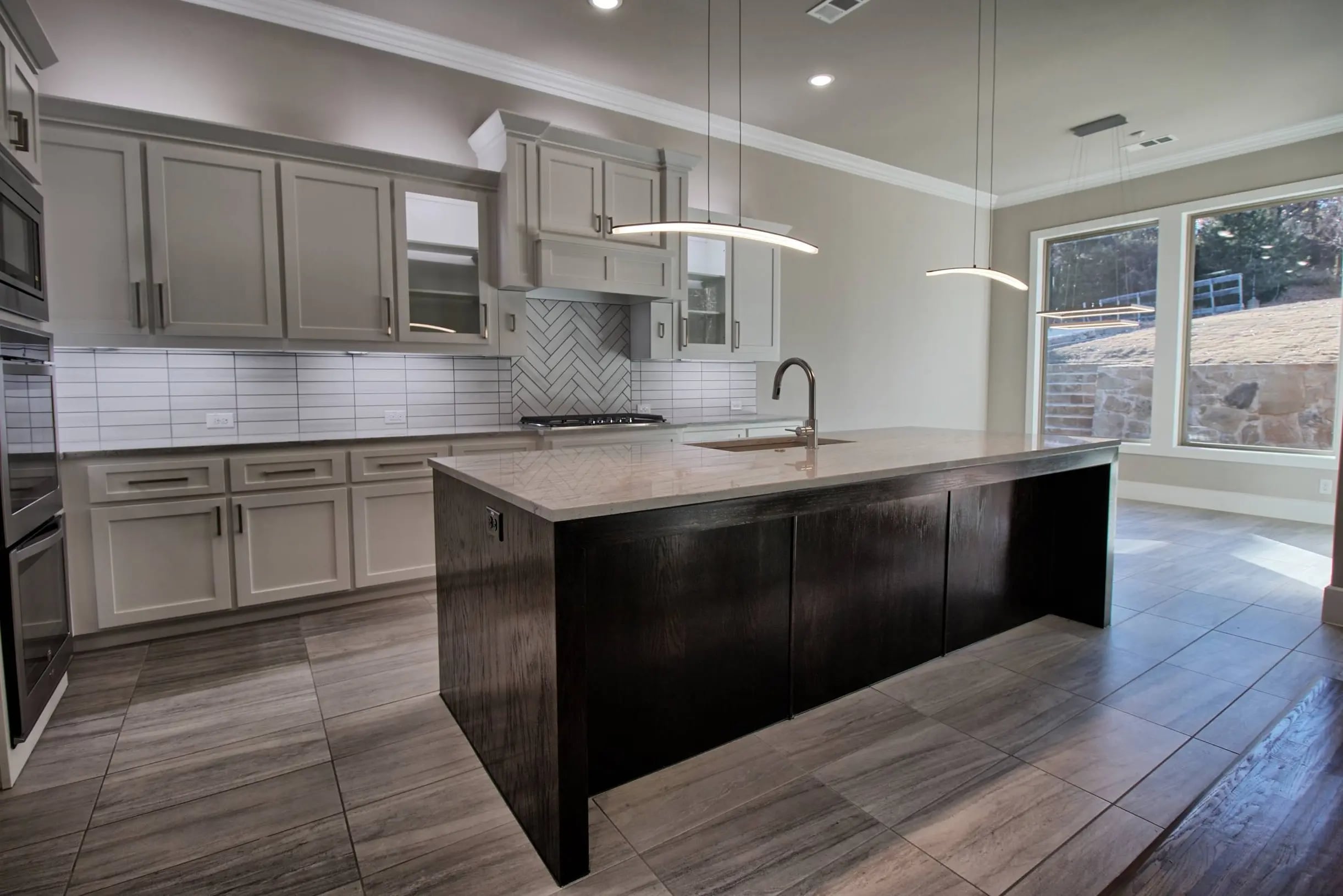 Contemporary kitchen with light gray shaker cabinets, herringbone tile backsplash, large picture windows, and a dark wood island with quartz countertop and built-in sink. Features modern pendant lighting and large format tile flooring.